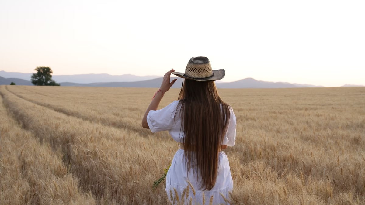A woman stands in a wheat field adjusting her straw hat, back turned, during a serene sunset.